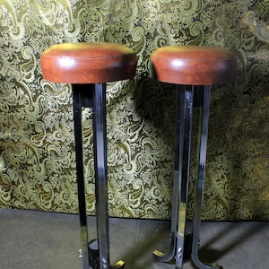 Op de afbeelding: Twee bruine leren barstoelen met chromen poten en een stervormige basis. De krukken staan voor een gouden, gepatroneerde achtergrond.
