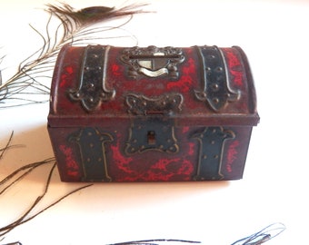 May include: A small, antique-style metal chest with a hinged lid. The chest is primarily burgundy with black metal accents and decorative flourishes. A small shield emblem is on the top. The chest has a keyhole and appears to be a vintage item.