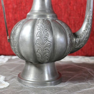 May include: A silver teapot with an ornate design and a handle. The teapot has a round body and a spout. The teapot is sitting on a white lace tablecloth.