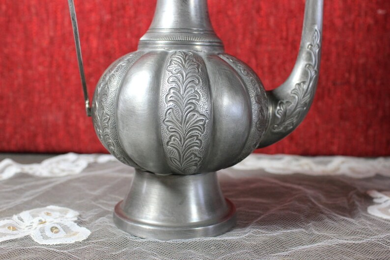 May include: A silver teapot with an ornate design and a handle. The teapot has a round body and a spout. The teapot is sitting on a white lace tablecloth.