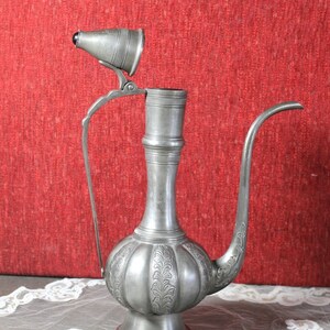May include: A silver teapot with an ornate design and a hinged lid. The teapot is sitting on a white lace tablecloth.