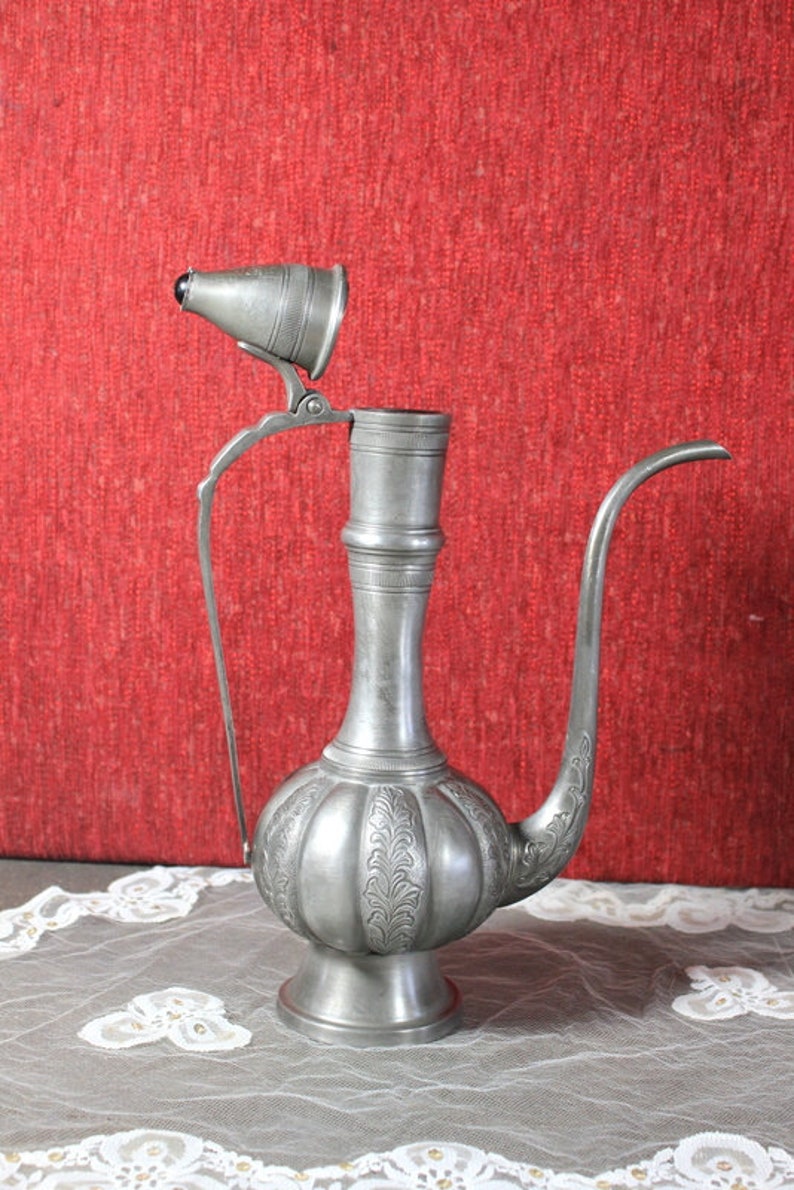 May include: A silver teapot with an ornate design and a hinged lid. The teapot is sitting on a white lace tablecloth.