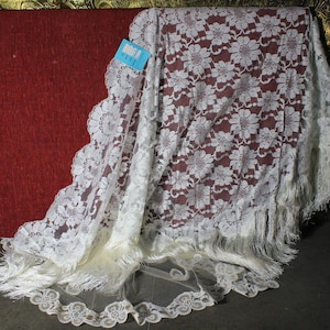 Può includere: Scialle di pizzo bianco con un motivo floreale e un bordo a frangia. Lo scialle è drappeggiato su una superficie rossa.