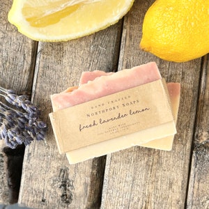Pode incluir: Duas barras de sabonete artesanal com um redemoinho rosa claro e branco, rotuladas como "fresh lavender lemon". O sabonete é embalado com uma faixa de papel castanho. Metade de um limão e raminhos de lavanda são visíveis.