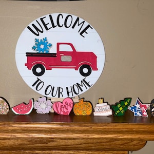May include: A wooden sign with the text "WELCOME TO OUR HOME" and a red truck carrying a blue snowflake. Below are various wooden cutouts including a flower, watermelon, heart, pumpkin, cake, tree, star, tombstone, and apple.