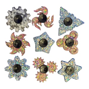 May include: A selection of glitter-covered fidget spinners in diverse shapes, such as gears, stars, and floral patterns. Each spinner features a central black button and is adorned with colourful glitter. These spinners are designed for stress relief and entertainment.