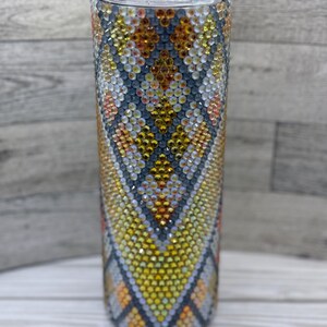 Puede incluir: Un vaso alto de acero inoxidable con un patrón geométrico de pedrería en tonos amarillos, naranjas y grises.