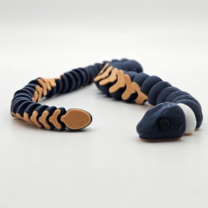 May include: A 3D printed articulated snake toy with a blue body, tan segmented scales, and a white eye. The snake is curled in a coil shape.