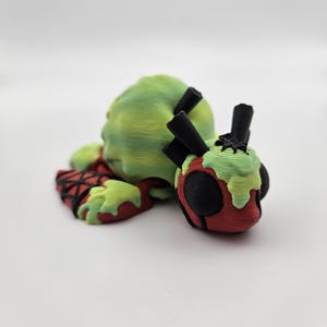 May include: A 3D-printed sculpture of a zombie-like figure with a green head and red and black clothing. The figure is lying down next to a smaller, similar figure with black antennae and green slime. The figures are on a white surface.