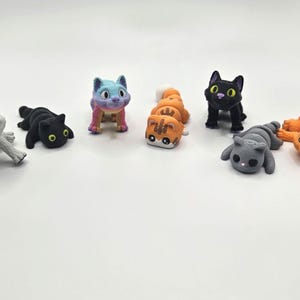 Op de afbeelding: Een verzameling kleine kattenbeeldjes in verschillende kleuren en poses. De katten zijn in tinten grijs, zwart, oranje en een regenboogverloop. Sommige staan, andere liggen. De beeldjes hebben grote ogen en staan tegen een witte achtergrond.