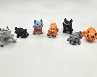 May include: A collection of small cat figurines in various colors and poses. The cats are in shades of gray, black, orange, and a rainbow gradient. Some are standing, while others are lying down. The figurines have large eyes and are set against a white background.