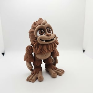Puede incluir: Una figura marrón de Bigfoot con una expresión amigable. La criatura de dibujos animados tiene ojos grandes, una barba peluda y pies detallados. La figura está de pie sobre una superficie blanca.