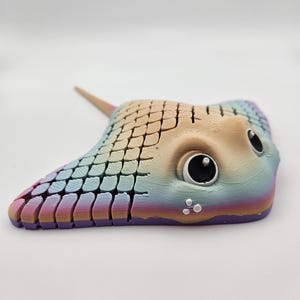 May include: A rainbow-coloured 3D printed stingray with a large eye and a small tail. The stingray is made of a textured plastic and has a purple underside.