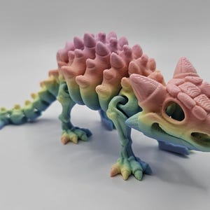 Puede incluir: Un modelo de esqueleto de dinosaurio impreso en 3D de color arcoíris. El modelo está en posición de caminar con la cabeza girada hacia un lado. El dinosaurio tiene una cola larga y una fila de púas en la espalda.