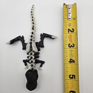 May include: A black and white plastic dinosaur skeleton model, approximately 6 inches long, with a white spine and black bones. The model is posed in a standing position with its tail raised.