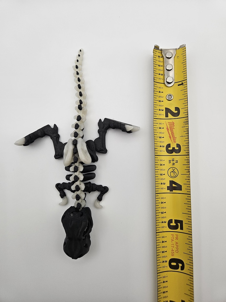 May include: A black and white plastic dinosaur skeleton model, approximately 6 inches long, with a white spine and black bones. The model is posed in a standing position with its tail raised.