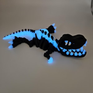 May include: A black glow-in-the-dark dinosaur skeleton figurine. The dinosaur is posed in a running position with its mouth open and teeth visible. The skeleton is made of plastic and has a smooth, glossy finish.