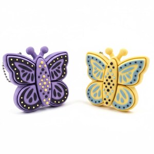 May include: Two butterfly-shaped keychains. One is purple with black and yellow accents, the other is yellow with blue accents. Both have a chain attached. The keychains are on a white background.