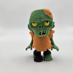 May include: A green and orange 3D printed zombie figurine with a missing chunk of its head. The zombie has yellow eyes and a toothy grin.