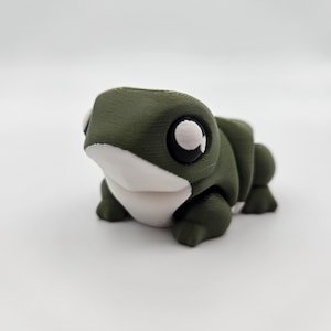 May include: A small, green 3D printed frog figurine with white eyes and a white belly.