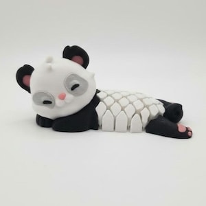 Peut inclure: Figurine de panda imprimée en 3D, en position détendue. Le panda a une tête et un corps blancs avec des accents noirs sur les oreilles, les pattes et autour des yeux. Le dos du panda est un motif géométrique blanc, avec des touches roses.
