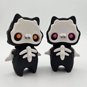 Puede incluir: Dos figuras en blanco y negro que se asemejan a esqueletos de gato. Las figuras tienen caras blancas con forma de calavera con ojos naranjas y rosas, capuchas negras y diseños de caja torácica blancos. Tienen pequeños apéndices blancos en forma de ala.