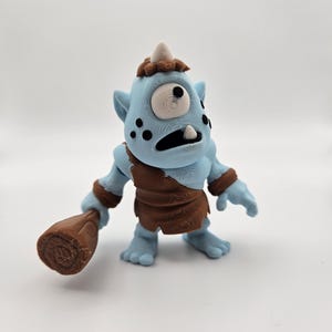 May include: A blue and brown one-eyed monster toy holding a brown club. The monster has a single white eye, a brown horn, and brown hair.