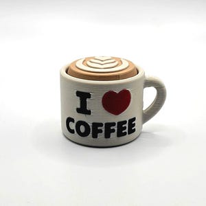 May include: A ceramic coffee mug with a handle, featuring the text "I ❤️ COFFEE" in black and a red heart. The mug is topped with a decorative, layered design resembling coffee art.