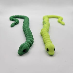 Puede incluir: Dos serpientes de juguete de plástico, una verde y otra amarilla, con ojos negros. Las serpientes están enroscadas en forma de 'S'.