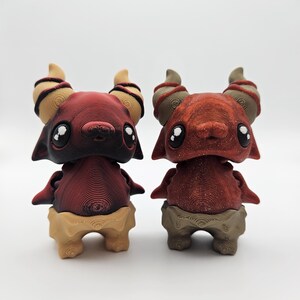 May include: Two small, red and brown, 3D printed figurines with horns and large eyes. The figurines are standing on a white surface.