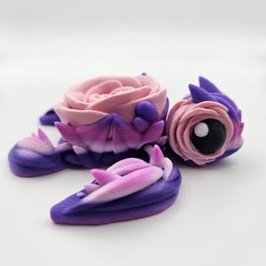 Könnte beinhalten: Eine kleine, lila und rosafarbene Schildkrötenfigur mit einer rosafarbenen Rose auf dem Rücken. Die Schildkröte hat ein schwarzes Auge und einen weißen Punkt in der Mitte.