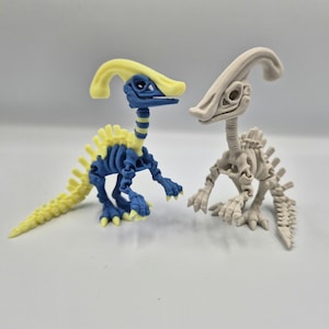 Puede incluir: Dos figuras de dinosaurios. Una es azul, amarilla y blanca, con un diseño de esqueleto. La otra es un esqueleto de dinosaurio beige. Ambas tienen un diseño de Parasaurolophus, con una cresta curva en la cabeza. Las figuras están sobre una superficie blanca.