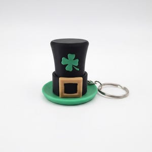 Può includere: Un portachiavi a tema San Patrizio. Presenta un cappello a cilindro nero in miniatura con un trifoglio verde, una fibbia beige e un bordo verde. Il portachiavi è attaccato a un anello di metallo argentato.
