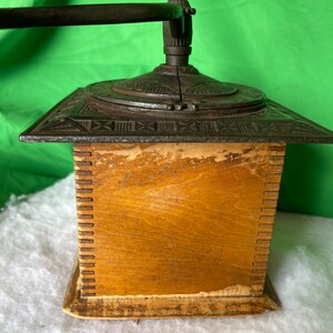 Op de afbeelding: Antiek gietijzeren koffiemolen met een houten basis. De molen heeft een kruk en een decoratief ontwerp aan de bovenkant.
