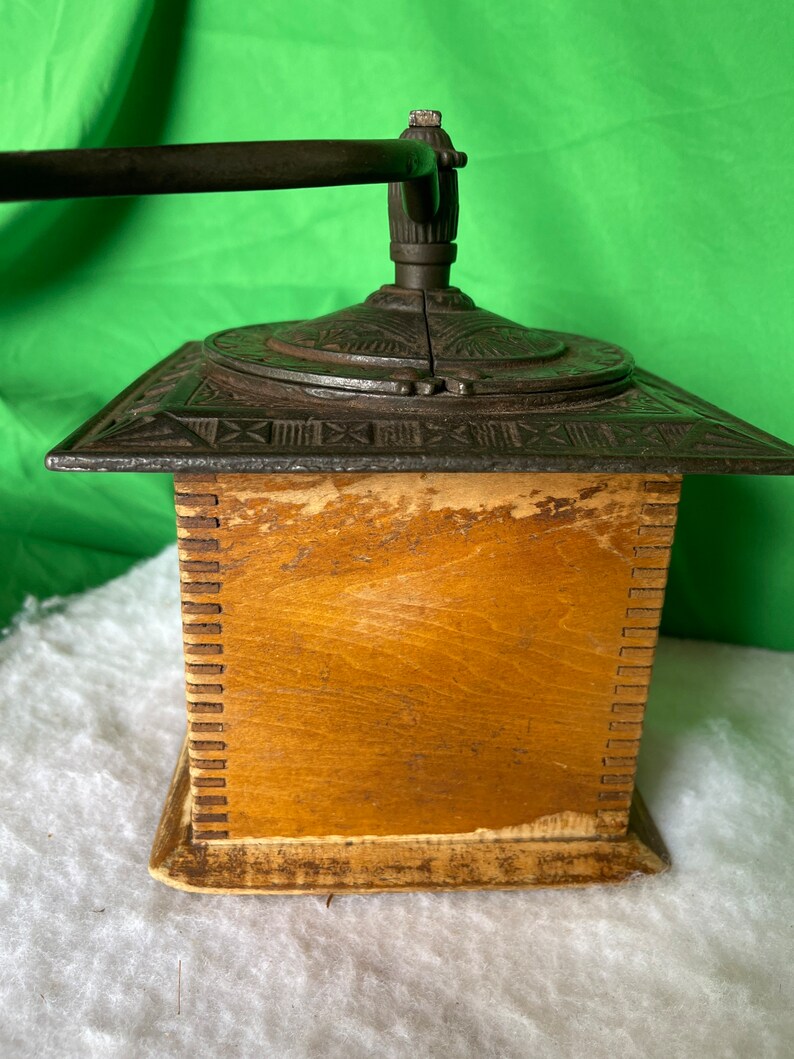 Op de afbeelding: Antiek gietijzeren koffiemolen met een houten basis. De molen heeft een kruk en een decoratief ontwerp aan de bovenkant.