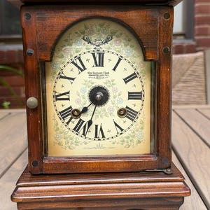May include: A wooden mantel clock with a dark brown finish, featuring an arched glass door and a cream-colored face. The dial displays black Roman numerals, floral accents, and the text New England Clock Farmington, Conn. U.S.A. 1974.