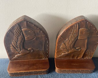 May include: A pair of brown leather bookends with a carved design of a bird in flight. The bookends have a wooden base and are decorated with metal studs.