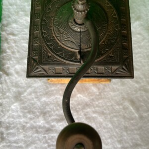 Op de afbeelding: Antiek gietijzeren koffiemolen met een decoratief ontwerp en een houten basis. De molen heeft een kruk en een metalen trechter.