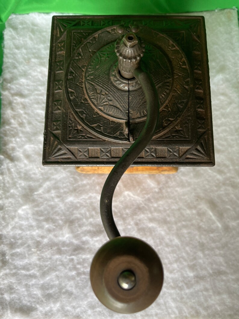 Op de afbeelding: Antiek gietijzeren koffiemolen met een decoratief ontwerp en een houten basis. De molen heeft een kruk en een metalen trechter.