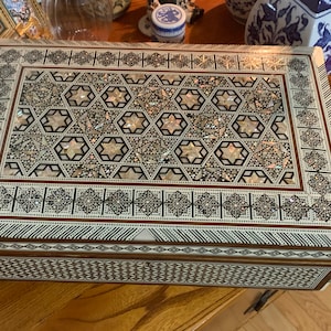 May include: A wooden box with a decorative inlay of mother-of-pearl. The box has a geometric pattern with a brown border.