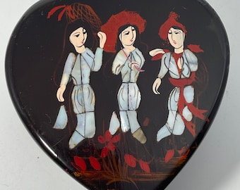 Op de afbeelding: Een hartvormige houten doos met een zwarte achtergrond en een geschilderd tafereel van drie vrouwen in traditionele kleding. De vrouwen zijn geschilderd in wit, rood en zwart met parelmoer accenten. De doos is versierd met rode en gouden accenten.