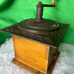 Op de afbeelding: Antiek gietijzeren koffiemolen met een houten basis en een handmatige slinger. De molen heeft ingewikkelde ontwerpen en een lade om de gemalen koffie op te vangen.