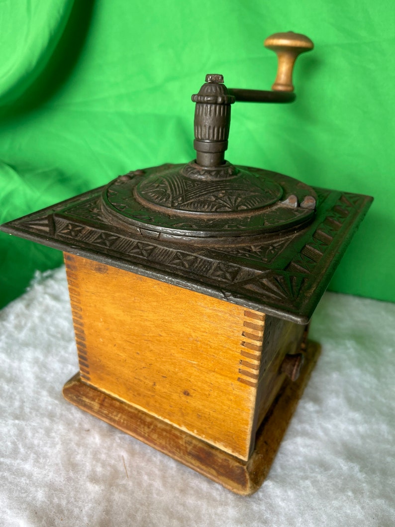 Op de afbeelding: Antiek gietijzeren koffiemolen met een houten basis en een handmatige slinger. De molen heeft ingewikkelde ontwerpen en een lade om de gemalen koffie op te vangen.
