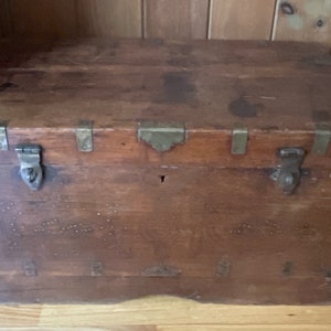 Puede incluir: Un cofre de madera vintage con herrajes de latón y una cerradura. El cofre tiene un aspecto desgastado y rústico y está decorado con pequeños remaches de metal.