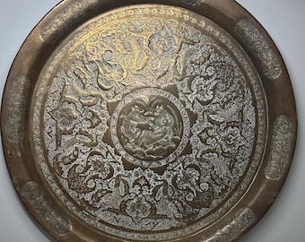 May include: A large, round, antique brass tray with intricate engraved patterns. The tray features a central medallion with figures and floral designs. The outer rim has a raised edge. The tray's color is a mix of gold and silver.