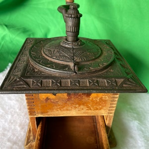 Op de afbeelding: Antiek gietijzeren koffiemolen met een houten handvat en een lade. De molen heeft een decoratief ontwerp op de bovenkant en de zijkanten.