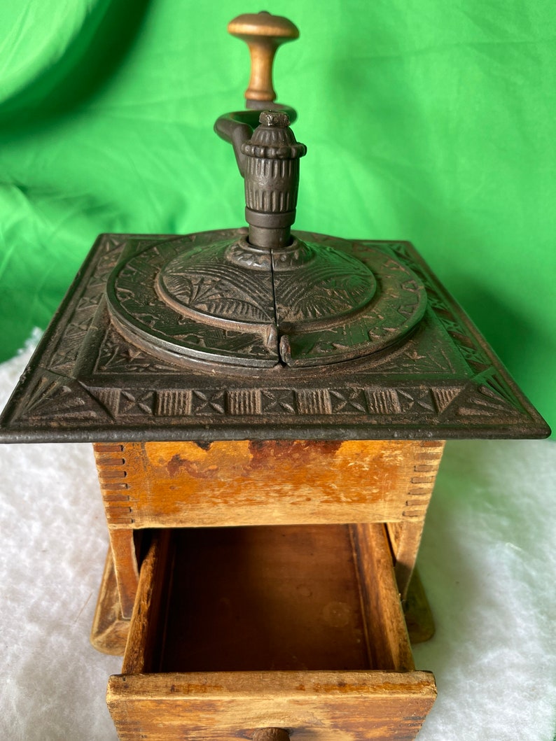 Op de afbeelding: Antiek gietijzeren koffiemolen met een houten handvat en een lade. De molen heeft een decoratief ontwerp op de bovenkant en de zijkanten.