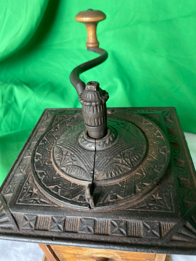 Op de afbeelding: Antiek gietijzeren koffiemolen met een houten kruk. De molen heeft een decoratief ontwerp aan de bovenkant en een lade eronder.