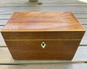 Op de afbeelding: Een houten doos met een lichtbruine afwerking en een klein sleutelgat aan de voorkant. De doos heeft een dunne, lichtkleurige rand rond de randen.