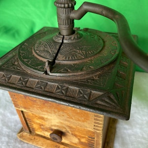 Op de afbeelding: Antiek gietijzeren koffiemolen met een houten basis en een lade. De molen heeft een decoratief ontwerp en een kruk.